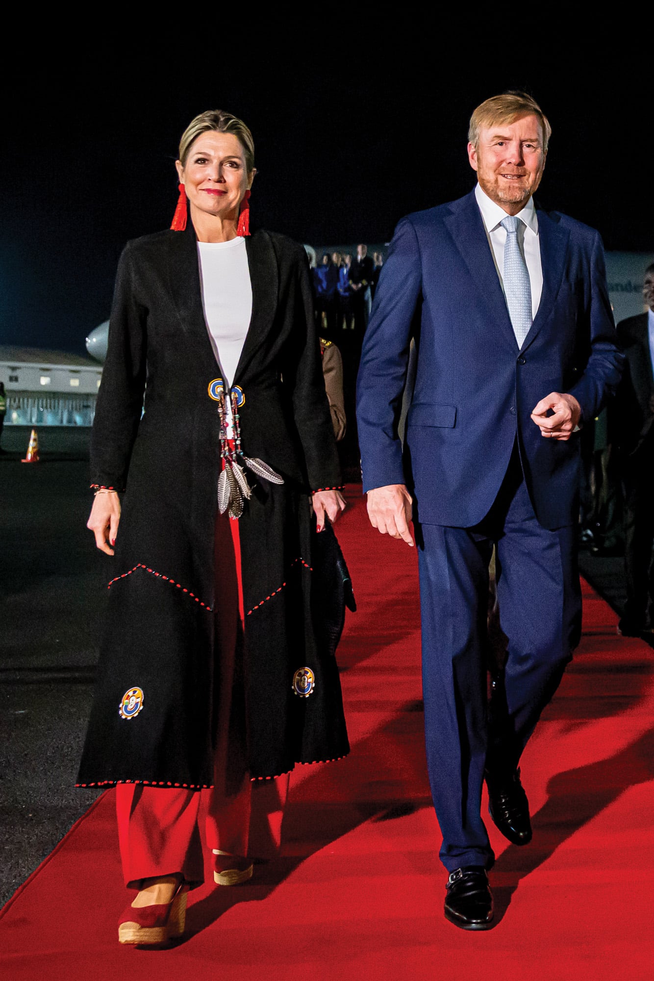 Para su primer día en Kenia, Máxima lució una blusa, pantalones anchos y un original abrigo con cuentas multicolores y plumas de la africana Anna Trzebinski. La Reina había comprado esa prenda, que está inspirada en las tradiciones de las tribus maasai y samburu, en 2007. Sumó, además, unos pendientes de cristales rojos con forma de borla, by Miccy.