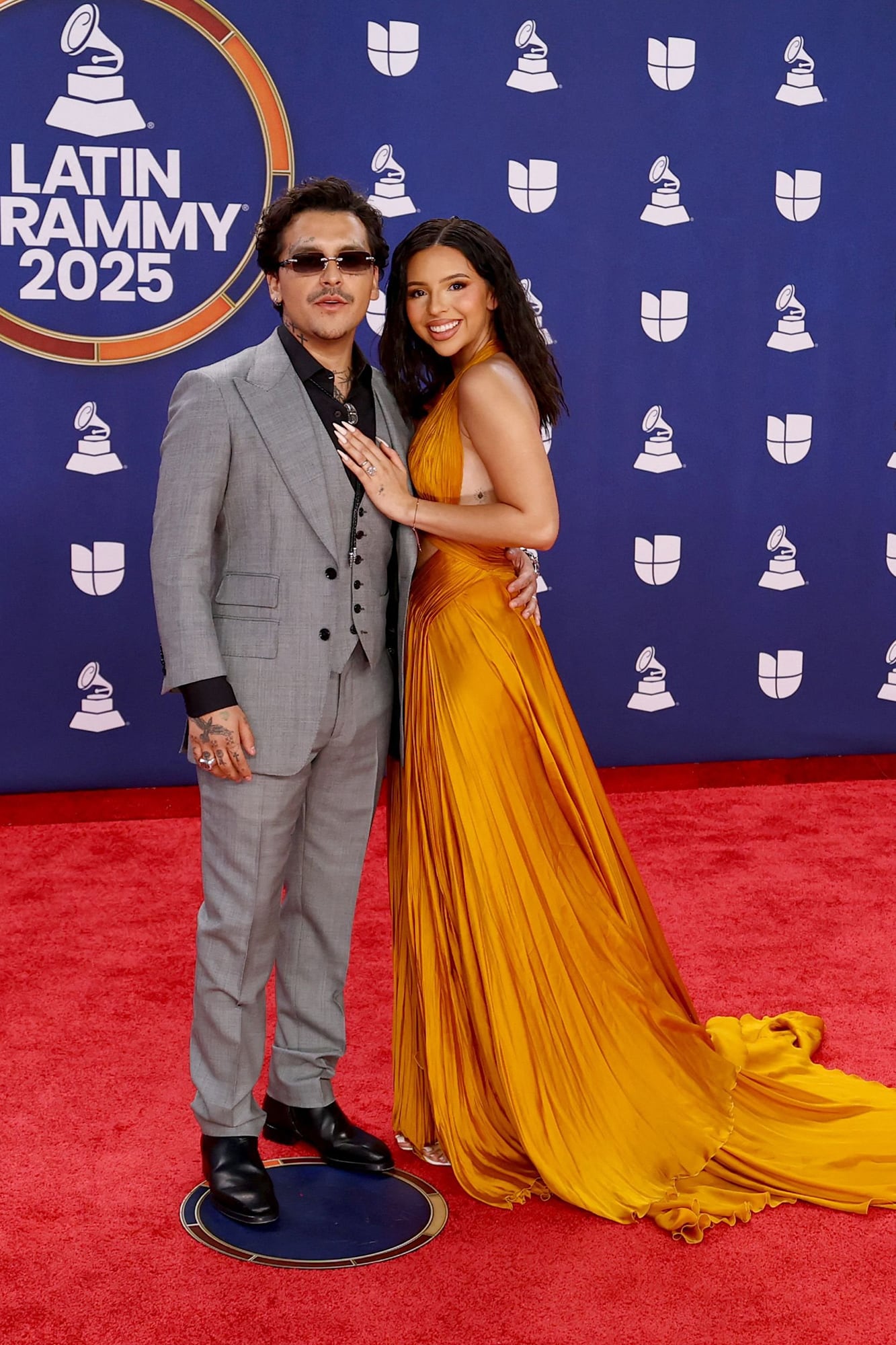 Christian Nodal llegó junto a Angela Aguilar. Mientras el cantante eligió un clásico traje de tres piezas, su pareja apostó por el color con este impactante modelo drapeado de escote halter 