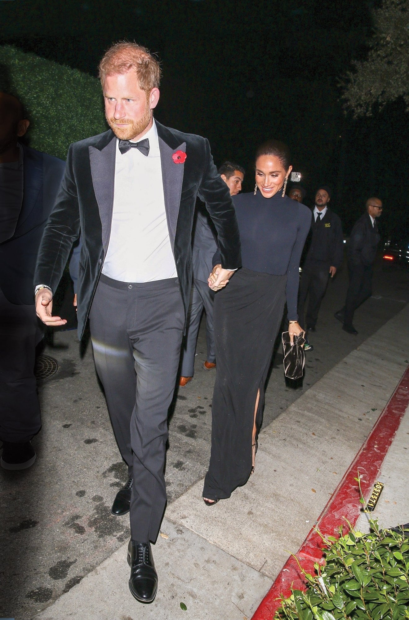 De impecable smoking, el príncipe Harry llega a la fiesta junto con Meghan Markle, con un vestido de cuello alto, mangas largas y drapeado a la altura de la cintura