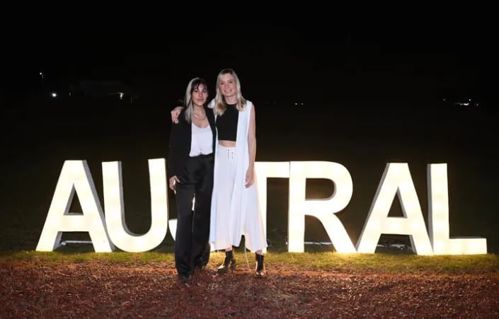 Delfina Lanusse y Chantal Leclercq posan juntas al aire libre, de noche, frente a un cartel luminoso de la Universidad Austral donde estudiaron juntas y se hicieron amigas íntimas