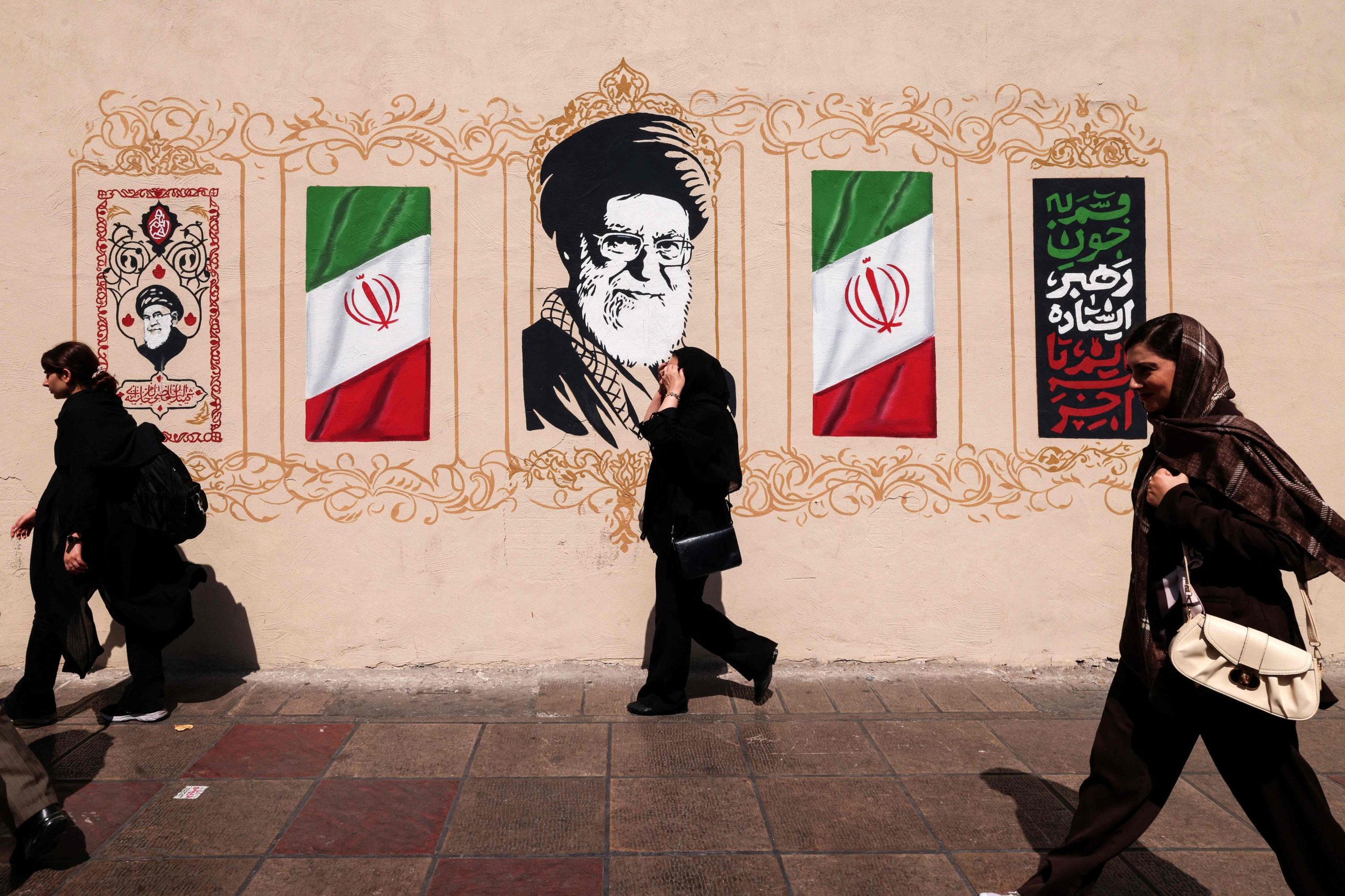 Iraníes caminan junto a un muro con una imagen del ayatollah Ali Khamenei, líder supremo de Irán asesinado, y la bandera nacional, en el centro de Teherán