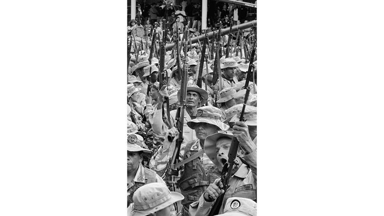Hombres con el uniforme de la Milicia Bolivariana levantan fusiles en alto durante un acto oficial. Entre rostros curtidos y boinas desajustadas, la disciplina del desfile se mezcla con gestos torpes y cuerpos desordenados