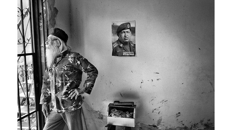 Un anciano con barba blanca y chaqueta de camuflaje observa desde la ventana. En la pared descascarada, un póster de Hugo Chávez lo vigila con la frase «Comandante Eterno»